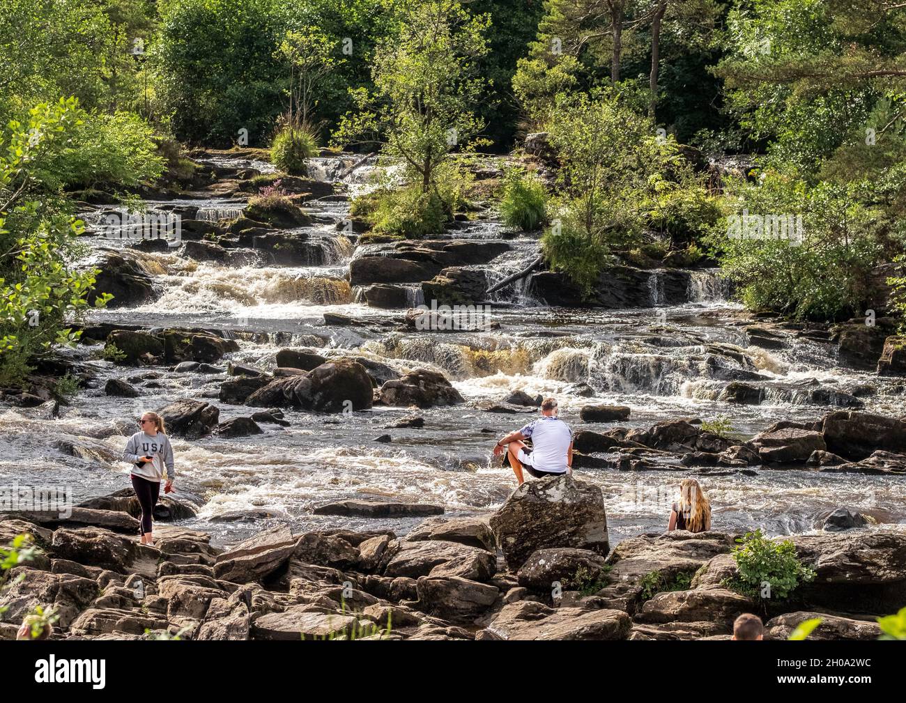 Visitatori seduti sulle rocce alle cascate di Dochart, sul fiume Dochart, appena fuori dal villaggio di Killin, Stirlingshire, Scozia Foto Stock