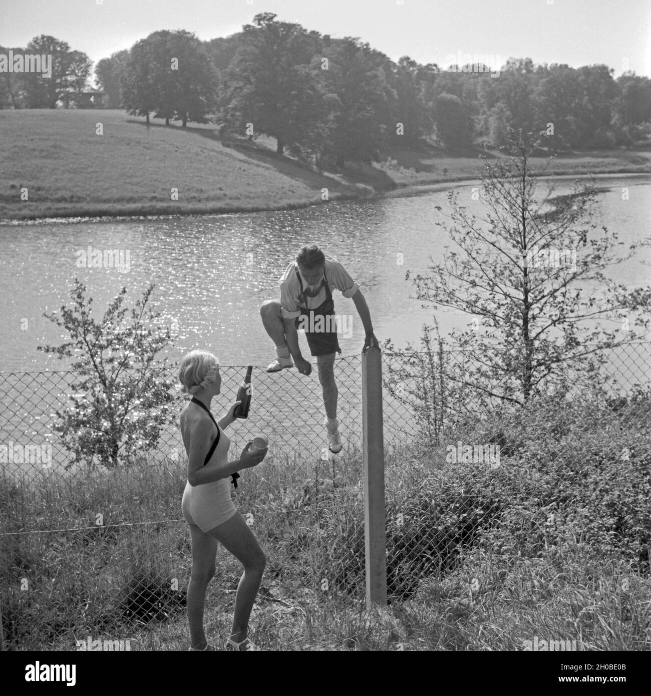 Ein junger Mann klettert über einen Zaun un einem vedere, eine Frau wartet mit einer Erfrischung, Deutschland 1930s. Un giovane uomo che si arrampica sul recinto, mentre la sua ragazza attende per lui con un drink, Germania 1930s. Foto Stock