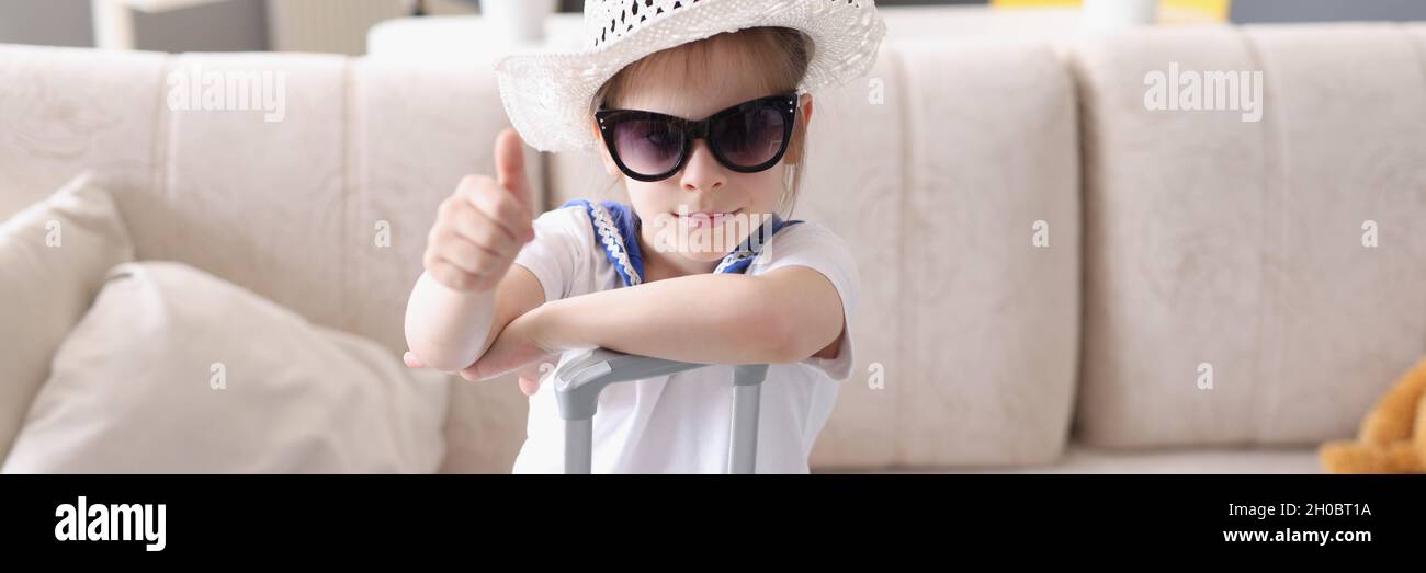 Bambina in cappello e occhiali da sole si alza con valigia e tiene i pollici in su Foto Stock