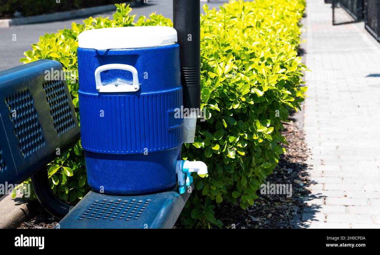 Primo piano di una caraffa d'acqua blu con tazze e una tazza di dispensatore seduto su una panca blu accanto ai campi da tennis circondati da cespugli. Foto Stock