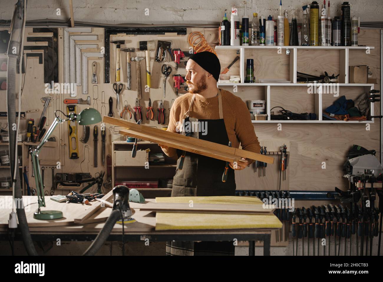Carpentiere in un cappello di orologio che tiene un pezzo di legno, in una  grande officina. Numero di attrezzi appesi alla parete Foto stock - Alamy