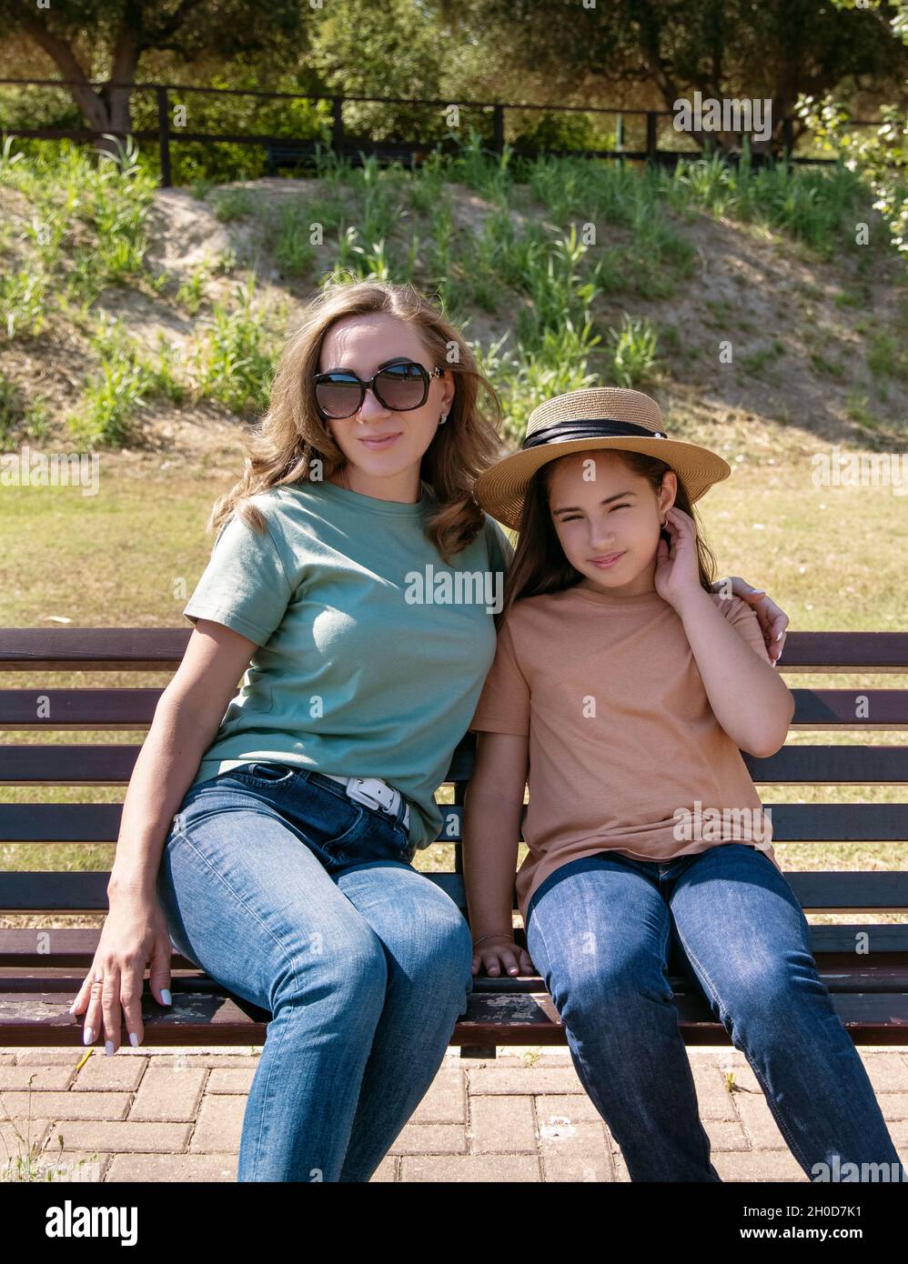 Madre e figlia seduta sulla panca di legno nel parco in giornata di sole. Una donna di mezza età e una bambina che indossa camicie e jeans. Mockup T-shirt. Foto Stock