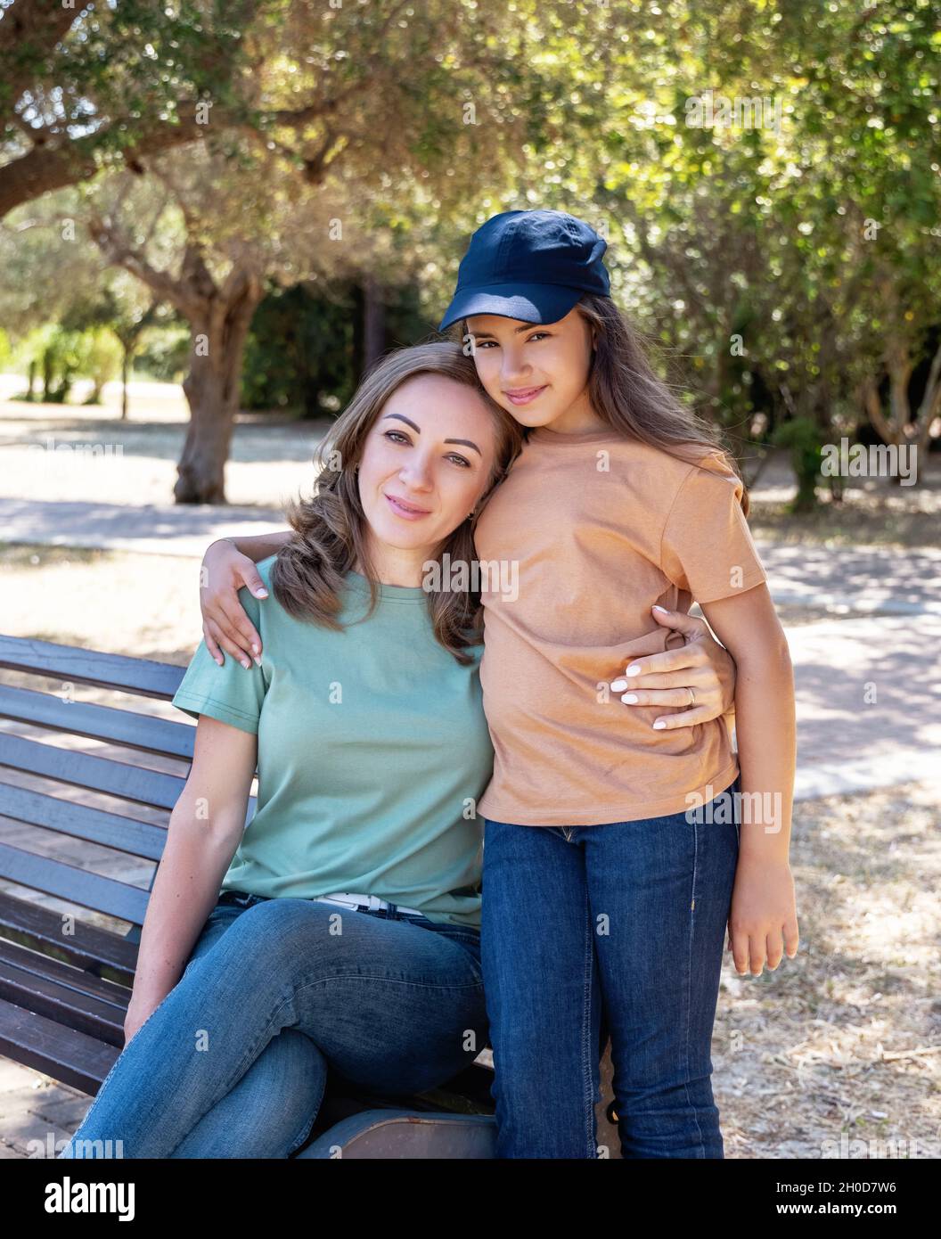 Madre e figlia seduta sulla panca di legno nel parco in giornata di sole. Una donna di mezza età e una bambina che indossa camicie e jeans. Mockup T-shirt. Foto Stock