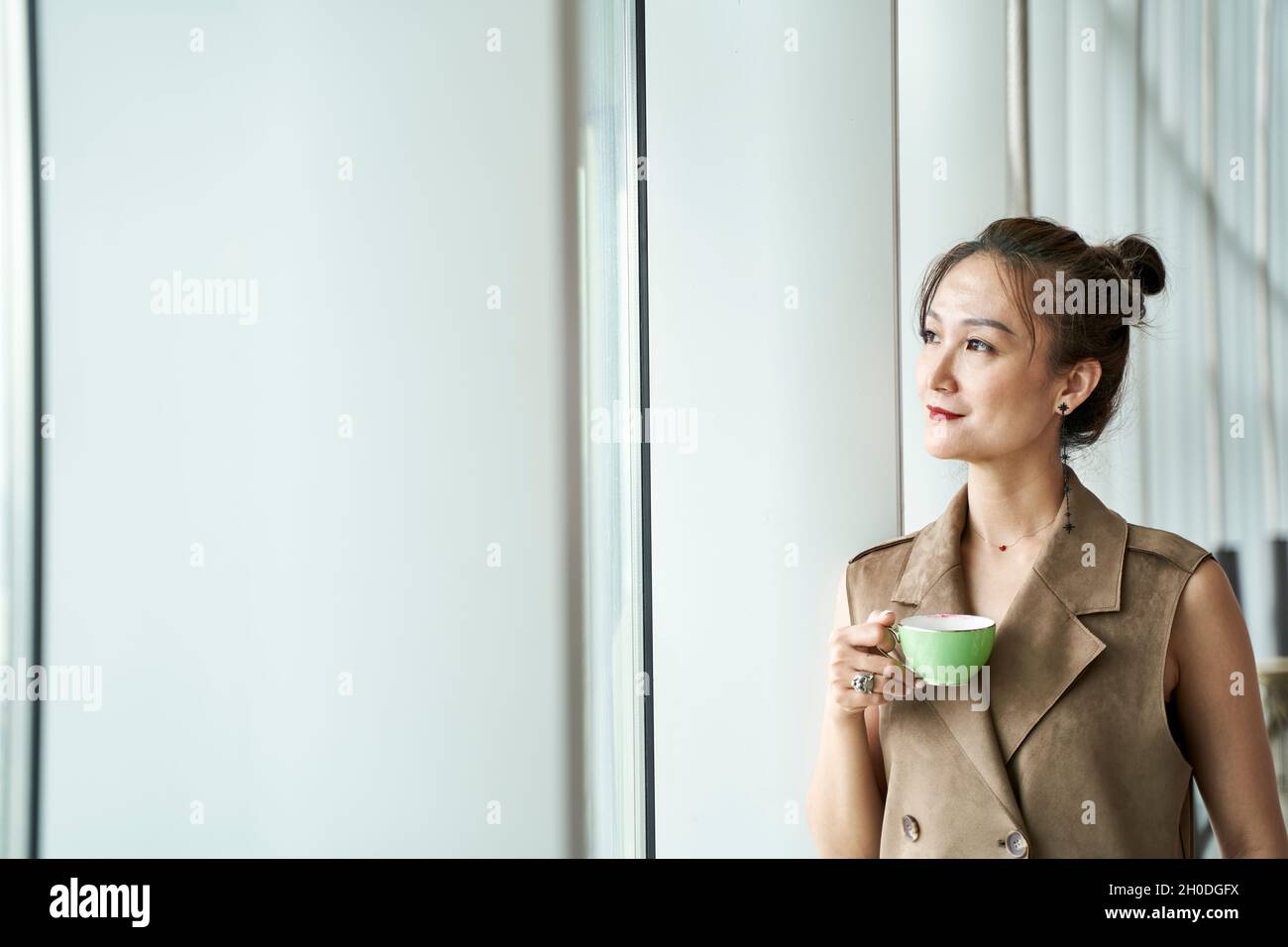 donna asiatica matura in piedi dalla finestra che guarda attraverso la finestra che tiene una tazza di caffè o tè Foto Stock