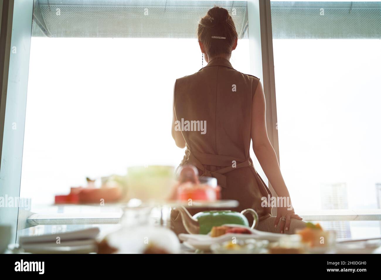 donna asiatica matura che guarda la vista attraverso la finestra della camera d'hotel con colazione sul tavolo Foto Stock