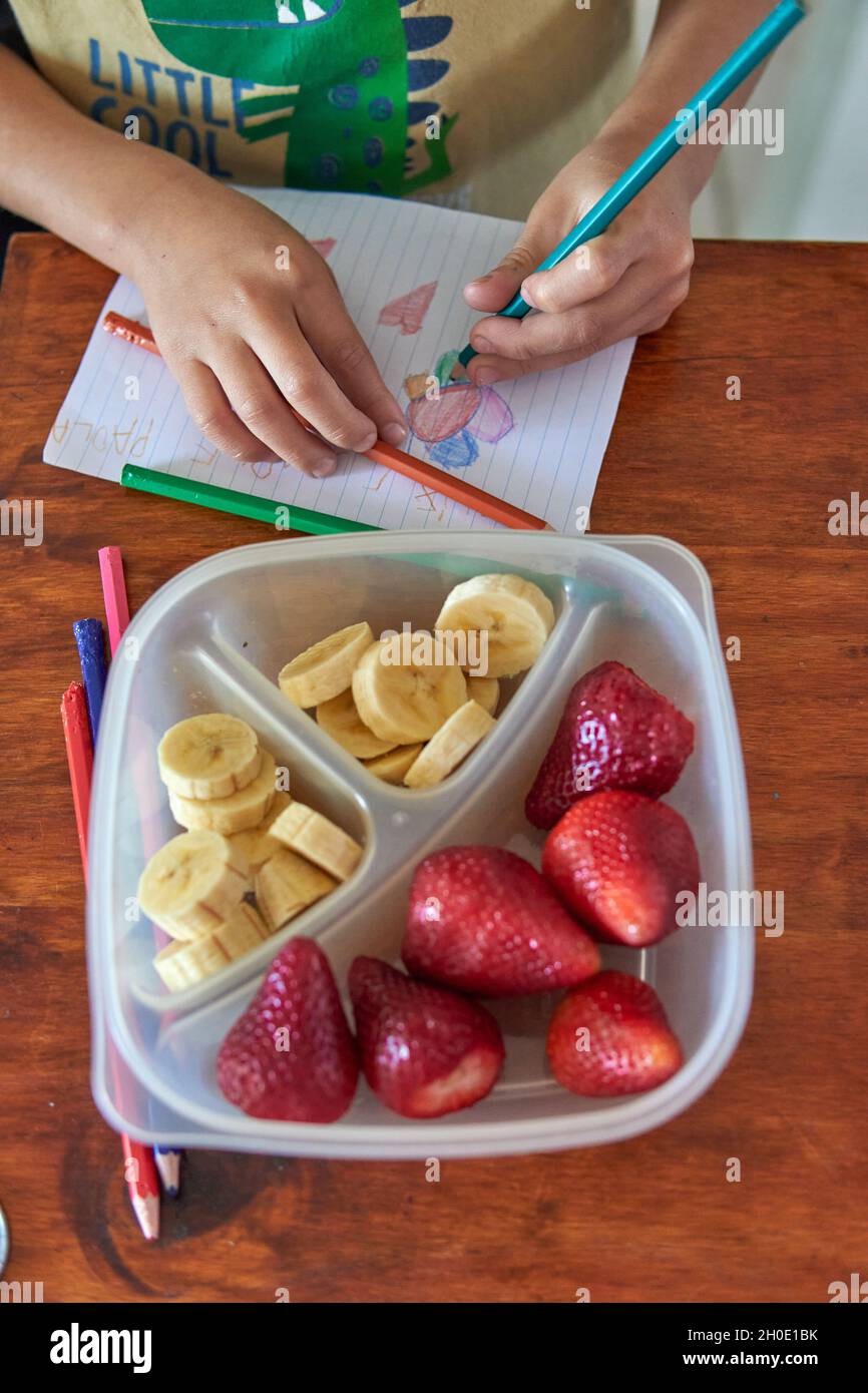 le mani del bambino disegnano con le pitture colorate mentre mangiano il suo spuntino sano di frutta fresca, banana e fragole. Verticale Foto Stock