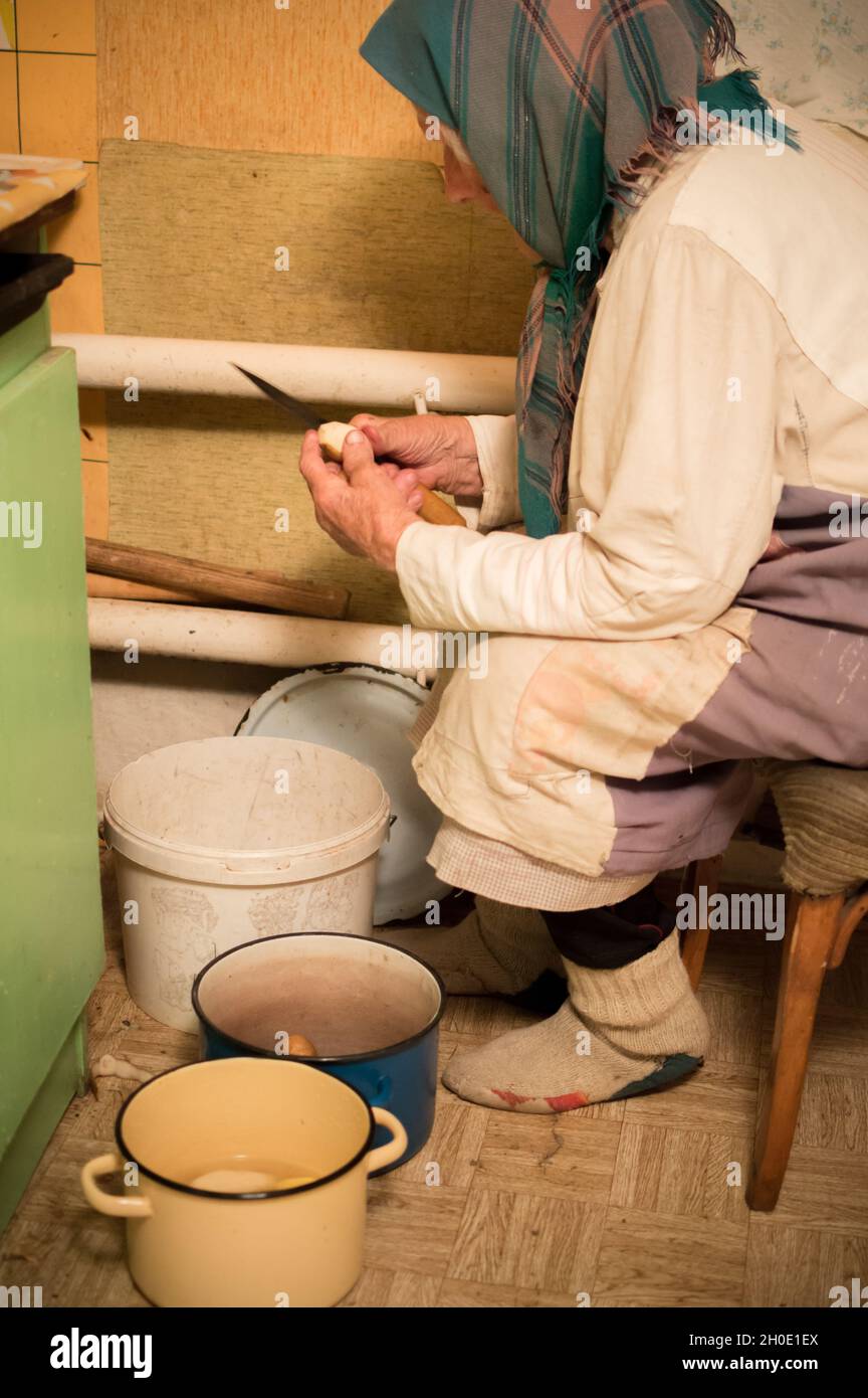 nonna di 90 anni in abiti sporchi spella le patate in una vecchia cucina Foto Stock