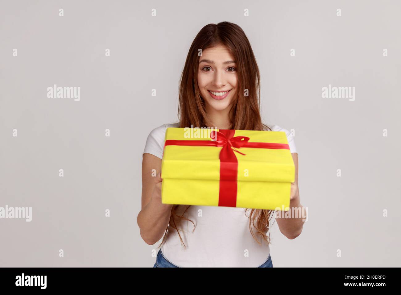 Ritratto di sorridente donna dai capelli scuri che dà giallo avvolto presente scatola, congratulandosi con vacanza, indossando bianco casual stile T-shirt. Studio interno girato isolato su sfondo grigio. Foto Stock