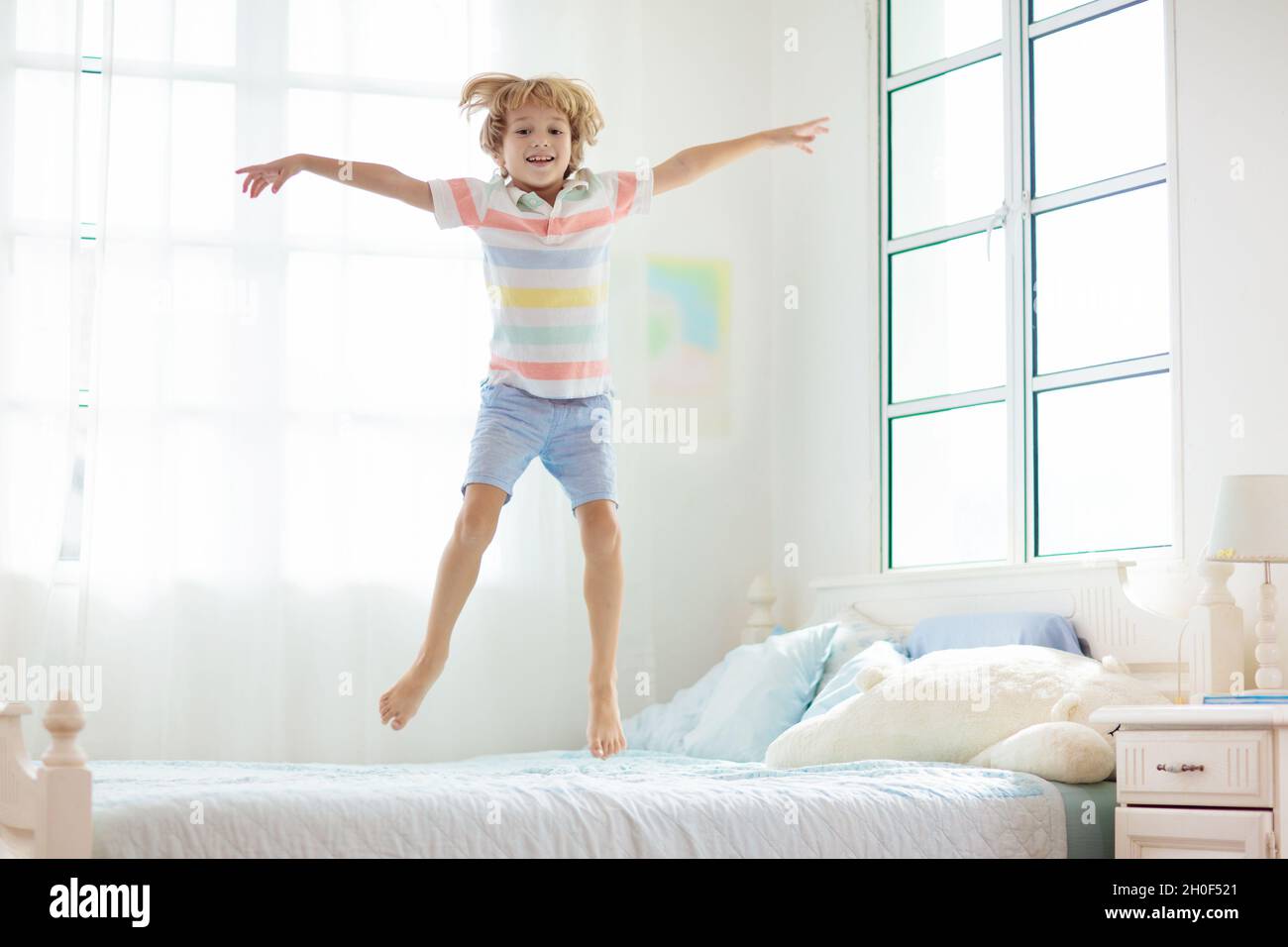Bambino che salta sul letto in camera bianca con grande finestra. I bambini  giocano a casa.