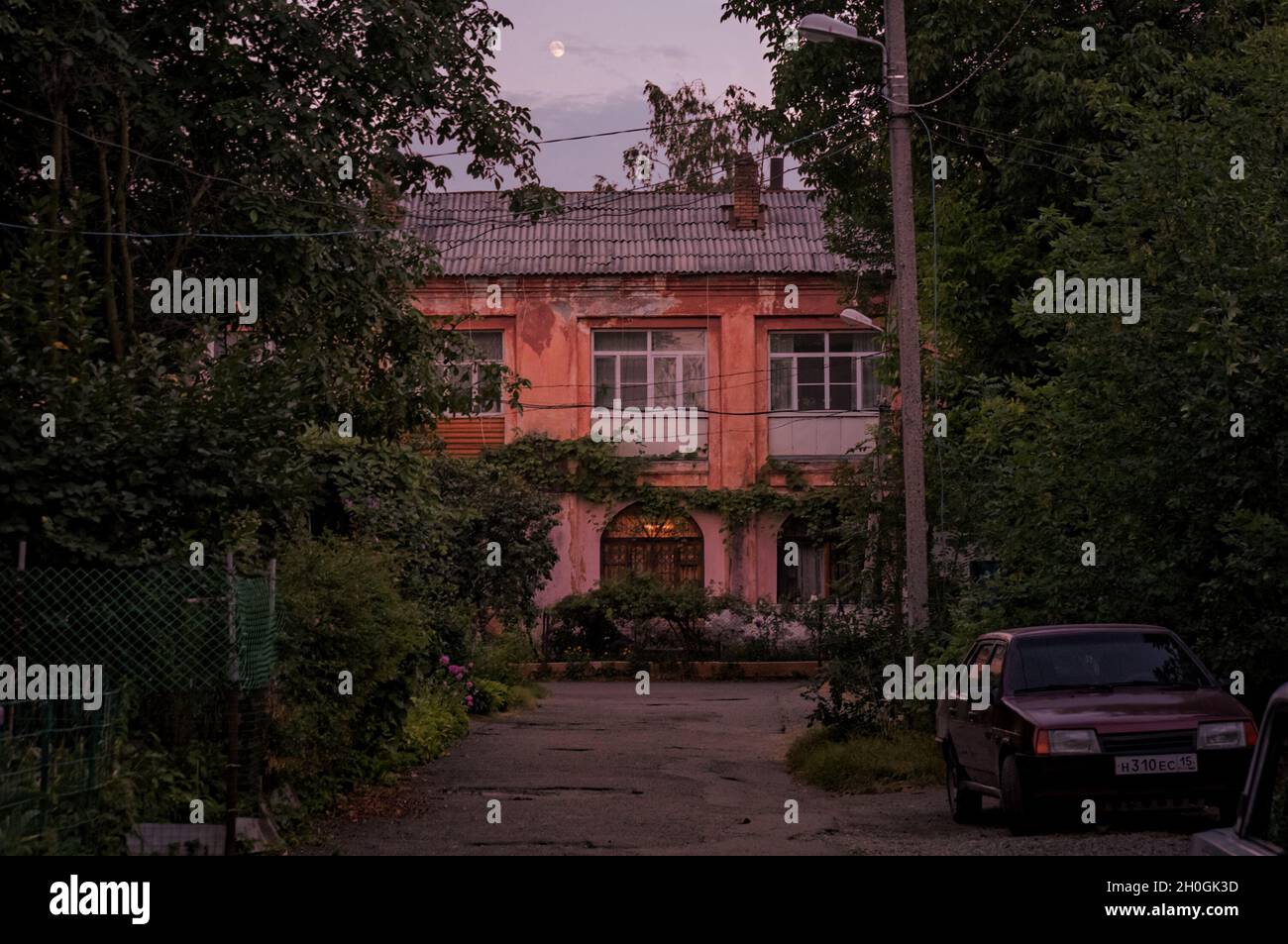 VLADIKAVKAZ, RUSSIA - 07 21 2021: Piccola città vecchia casa a due piani cortile annegato nel verde con una vecchia macchina LADA parcheggiata sulla strada nel crepuscolo Foto Stock