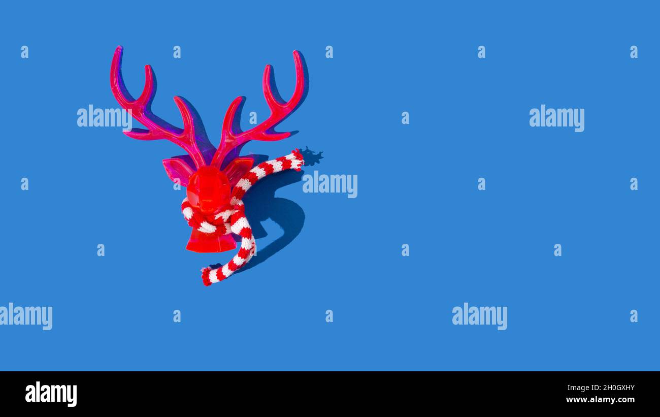Testa di renna rossa al neon con una sciarpa invernale a righe rossa e bianca isolata su sfondo blu. Concetto creativo di Natale e Capodanno. Foto Stock
