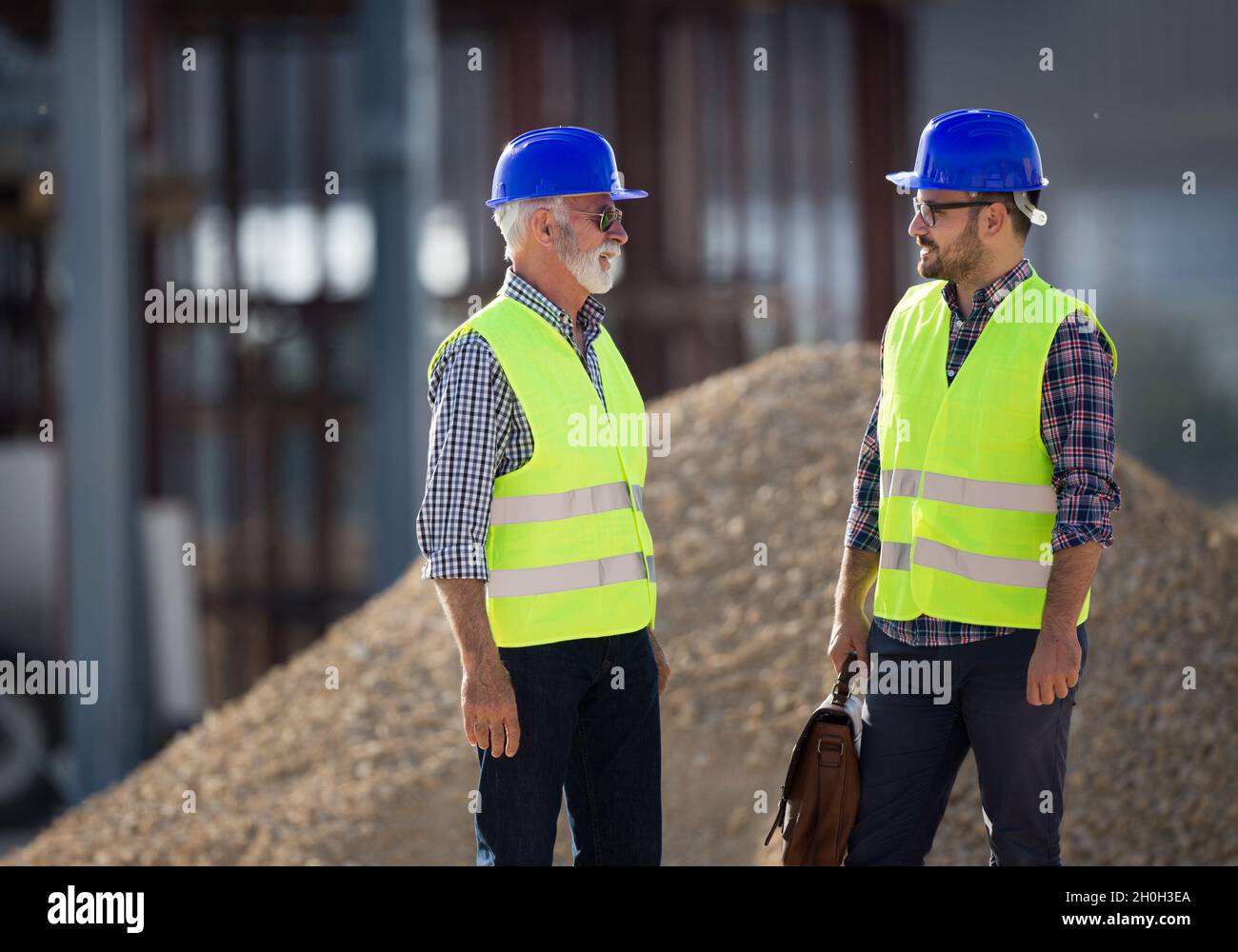 Due ingegneri con caschi e giubbotti che parlano in cantiere di fronte alla costruzione in metallo Foto Stock