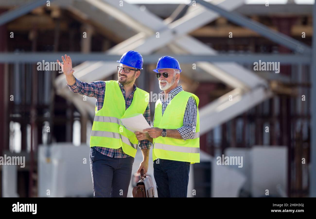 Due ingegneri con caschi e giubbotti che parlano in cantiere di fronte alla costruzione in metallo Foto Stock