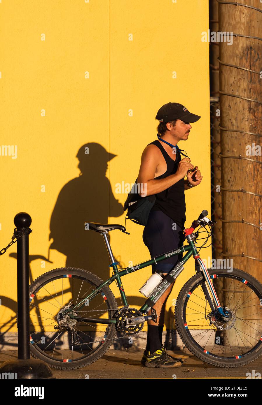 Un uomo che cammina sul marciapiede con la sua bicicletta e la sua ombra sul muro giallo di un edificio. Luci e ombre che suonano durante il tramonto. Vancouver, British Columbia Foto Stock