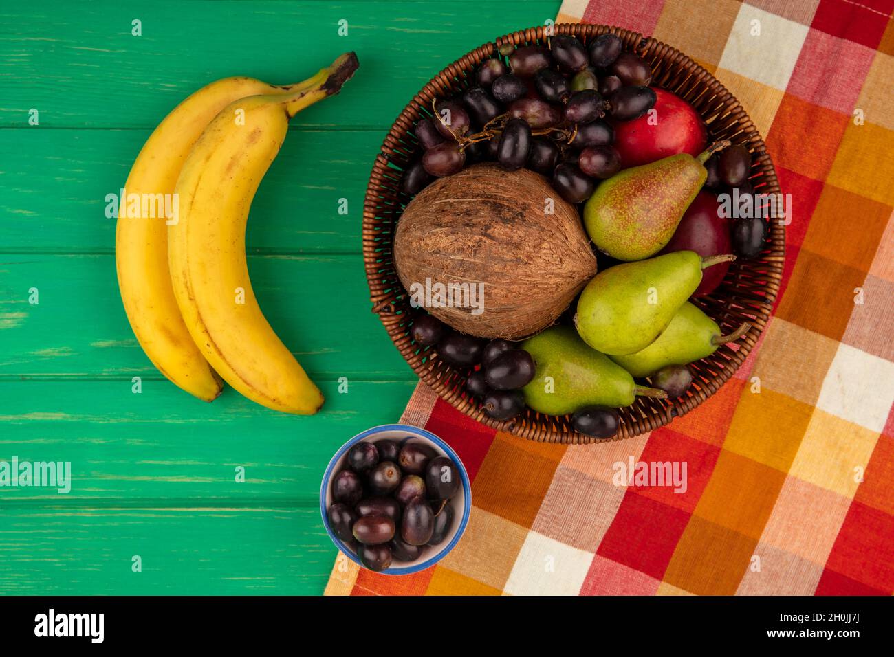 vista dall'alto di frutta come pera pesca uva cocco in cestino su tela di plaid e banane su sfondo verde Foto Stock