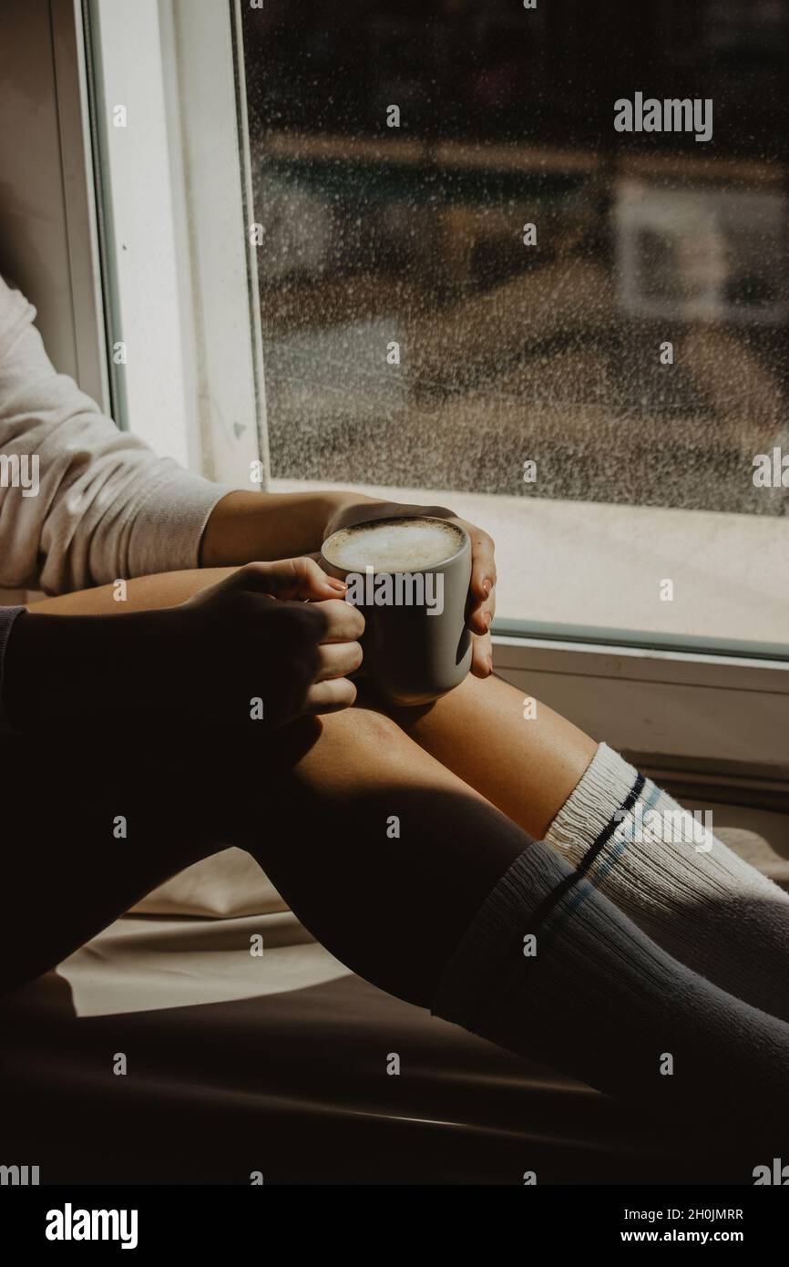 Una donna irriconoscibile alla finestra della sua casa seduta con un caffè caldo latte mentre si gode il tempo con se stessa. Fotografia del caffè di Still Life. Foto Stock