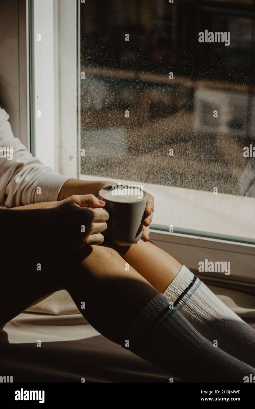Una donna irriconoscibile alla finestra della sua casa seduta con un caffè caldo latte mentre si gode il tempo con se stessa. Fotografia del caffè di Still Life. Foto Stock