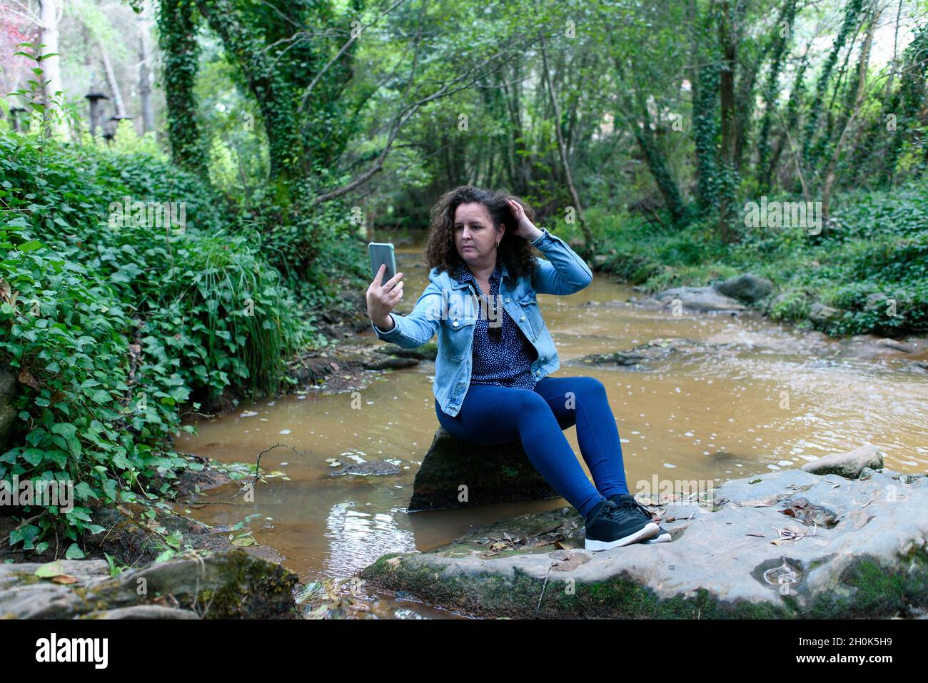 Donna seduta su una roccia accanto a un ruscello che scorre attraverso una foresta di gallerie, prende un selfie con il suo smartphone, tenendo i capelli in mano. . Foto Stock