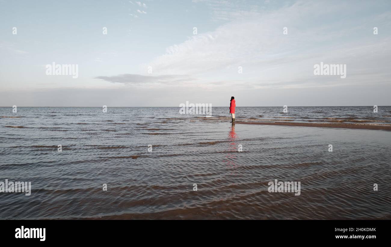 Una donna in giacca rossa guarda le onde schiumose. Ventoso giorno d'autunno alla riva del lago Ilmen. Foto Stock