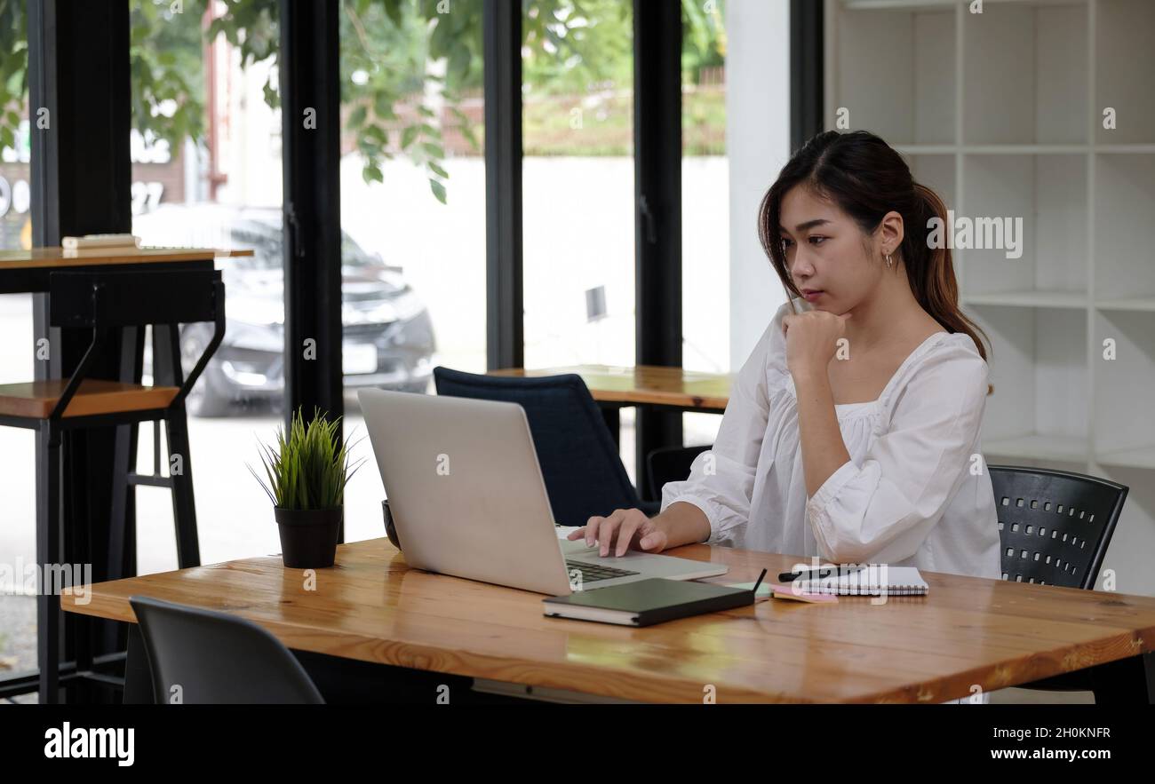 Donna di affari asiatica focalizzata che lavora in ufficio studiando in linea guardando il laptop, il dipendente serio o lo studente che guardano le informazioni di scrittura di webinar in Foto Stock