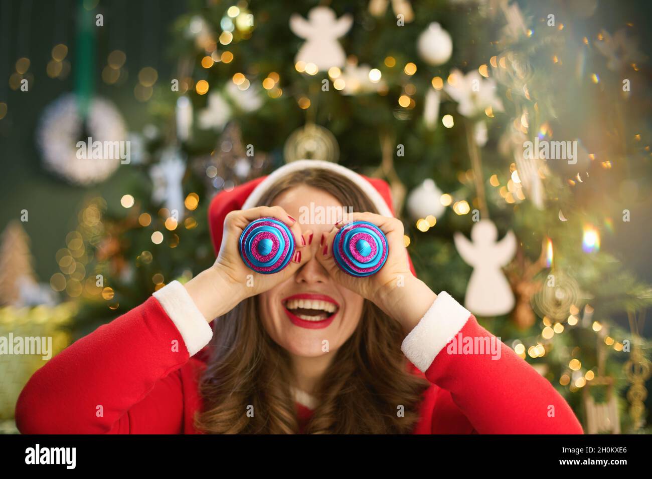 Ora di Natale. Sorridente casalinga alla moda con cappello di Santa usando le palle di Natale come occhi vicino albero di Natale nella casa moderna. Foto Stock