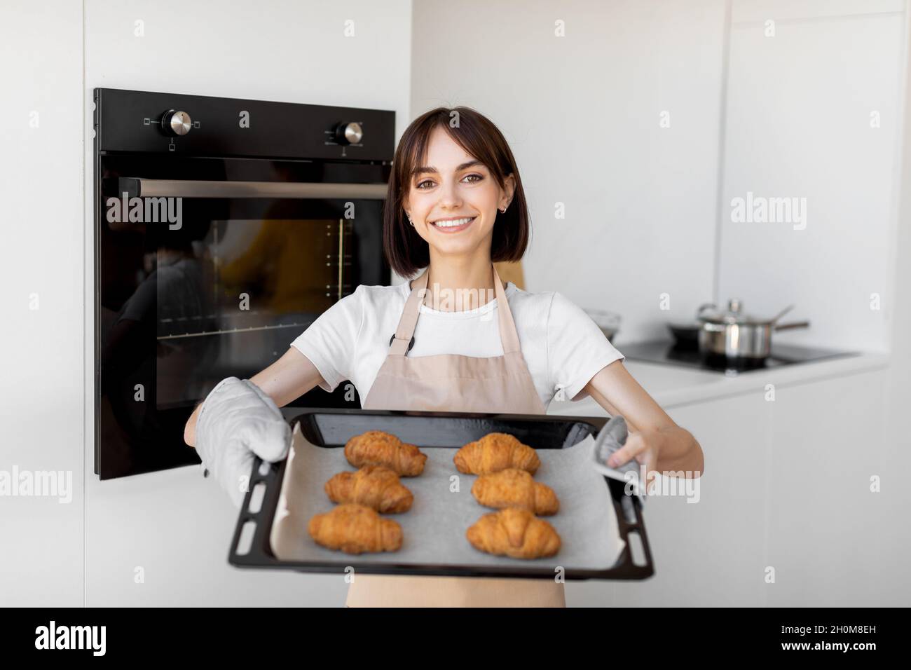Felice giovane casalinga che tiene vassoio con cornetti appena sfornati, in piedi in cucina e sorridendo alla macchina fotografica Foto Stock