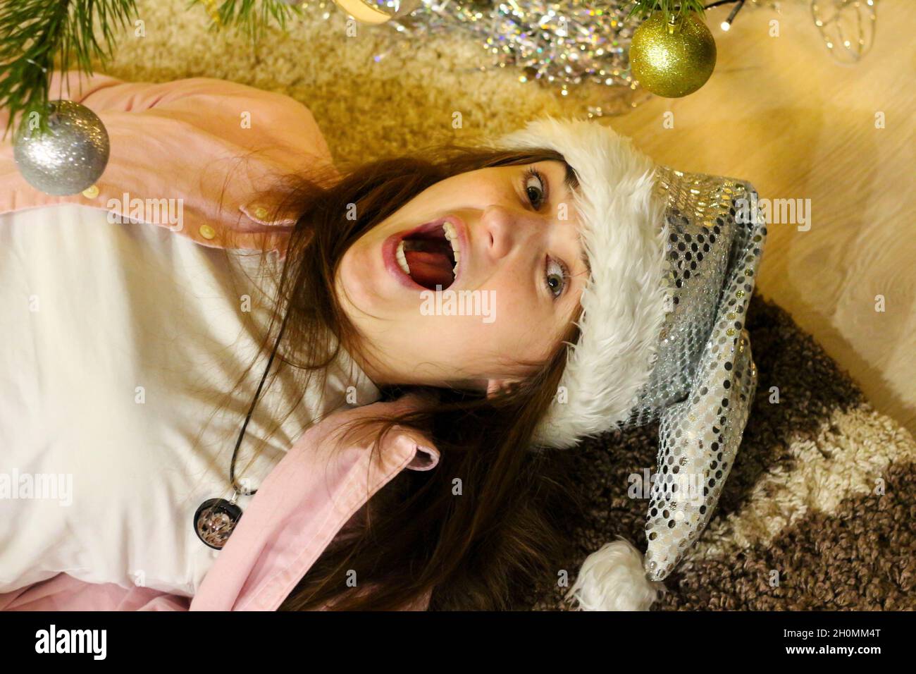 Ritratto di pazzo sorpreso giovane donna brunetta 20-25 anni indossando bianco santa cappello e camicia rosa. Sdraiato su tappeto sotto l'albero di Natale sullo sfondo. Foto Stock