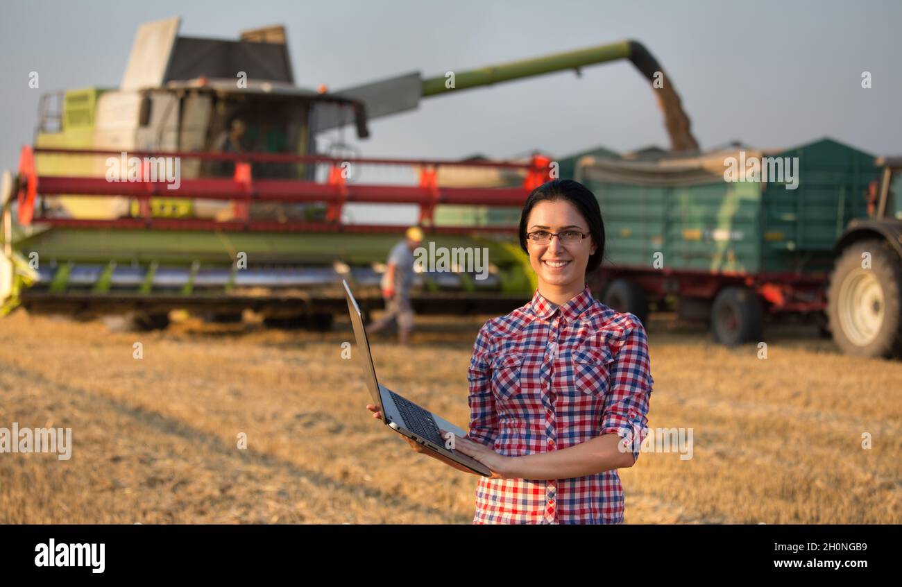 Bella giovane donna d'affari con un computer portatile davanti alla macchina della mietitrebbia e al trattore con rimorchio durante la mietitura del grano Foto Stock