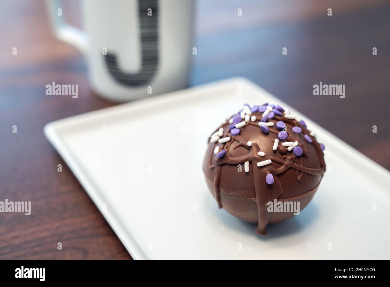 Una sfera o sfera calda della bomba di cacao ricoperta di cioccolato marrone gocciolato e spolverini viola e bianchi posti su un piatto da portata bianco con monogrammo J coff Foto Stock