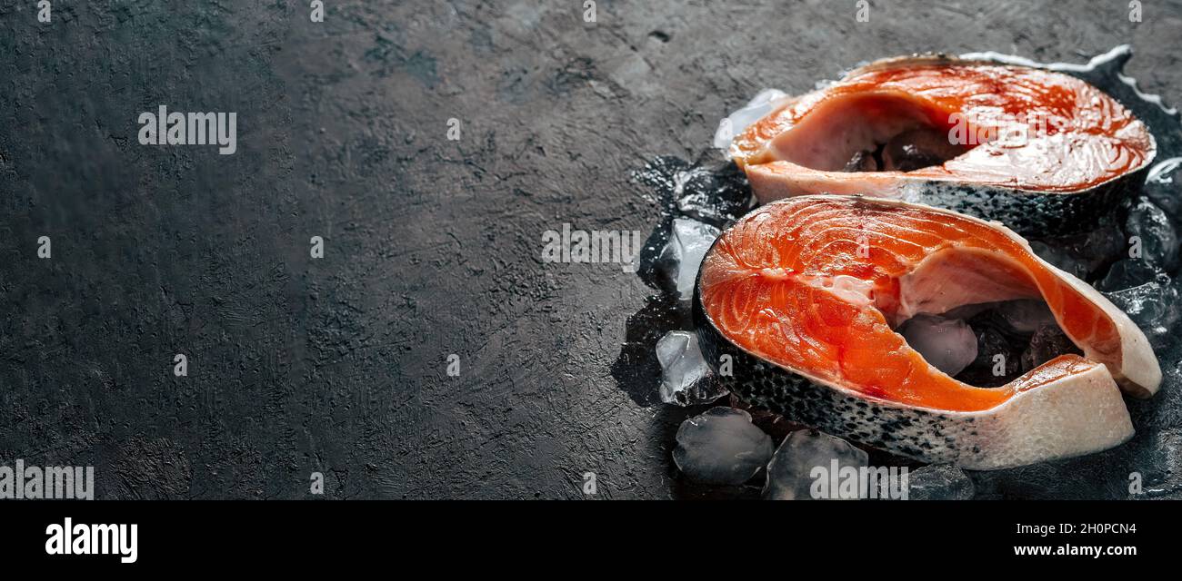 Due bistecche di salmone selvatico crudo su ghiaccio su sfondo nero testurizzato. Bistecche di salmone con spazio di copia a sinistra. Banner orizzontale lungo Foto Stock