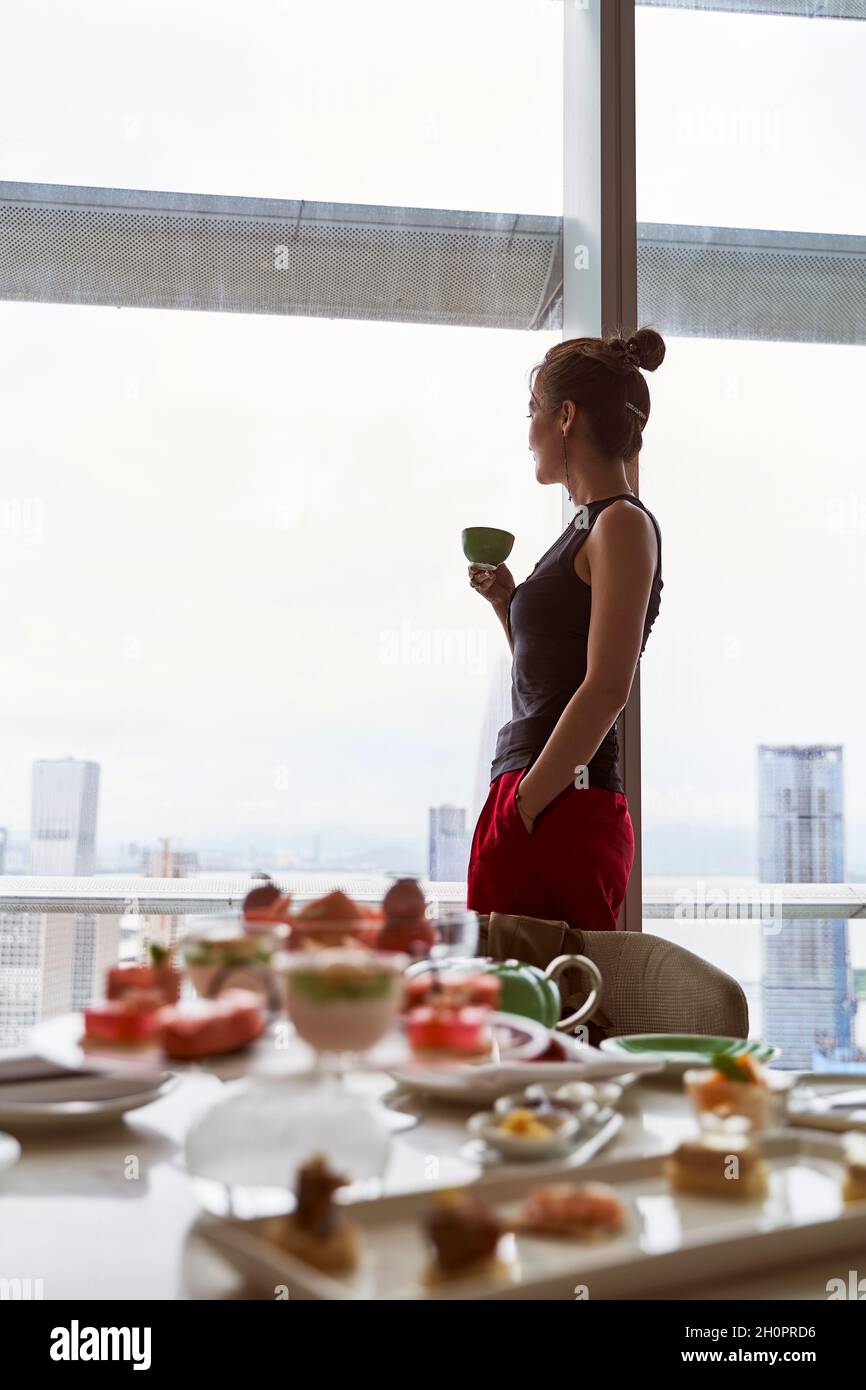 donna asiatica matura in piedi accanto alla finestra che guarda la vista della città con cibo sul tavolo in una camera d'hotel Foto Stock