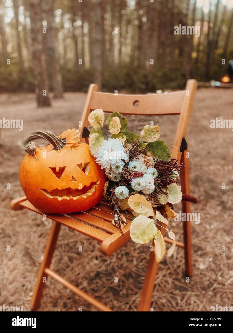 Decorazioni di Halloween. Cric-o-lanterna di zucca arancione intagliata con faccia spoky e bouquet di fiori autunnali su sedia di legno nella foresta Foto Stock