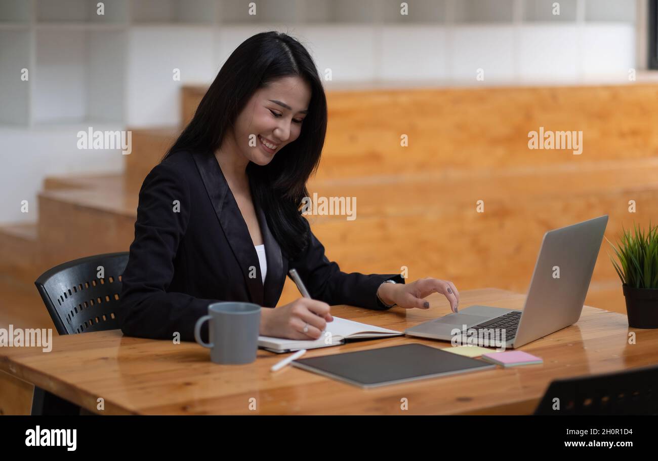 Donna d'affari asiatica che lavora su un computer portatile durante la presa di appunti in ufficio. Concetto di webinar per riunioni online. Foto Stock