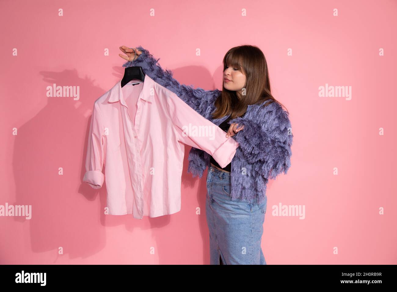giovane donna stilista di moda tiene appendiabiti con camicia su sfondo rosa in studio. concetto di guardaroba di base. Blogger insegna le lezioni sulla creazione di stile Foto Stock