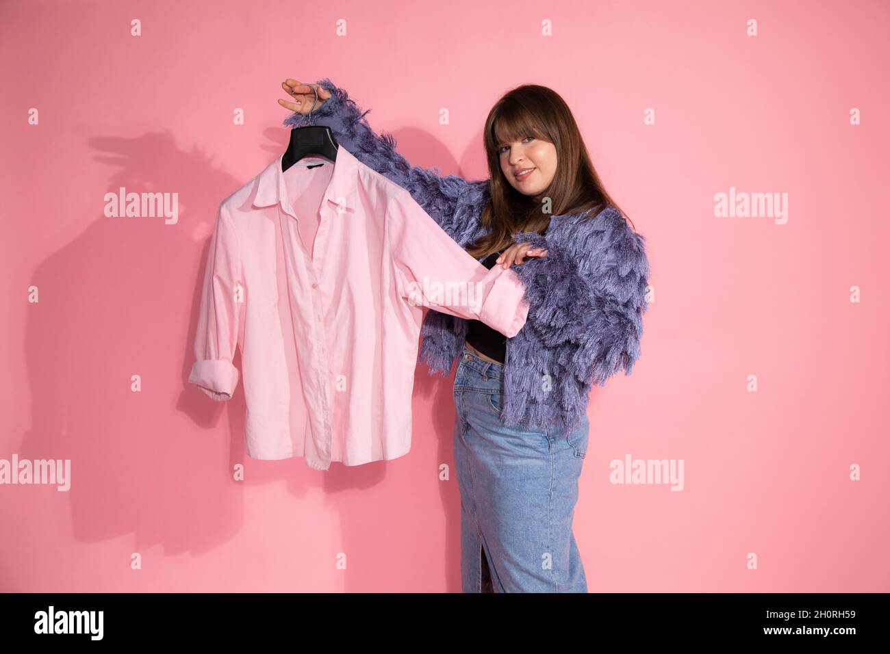 giovane donna stilista di moda tiene appendiabiti con camicia su sfondo rosa in studio. concetto di guardaroba di base. Foto Stock