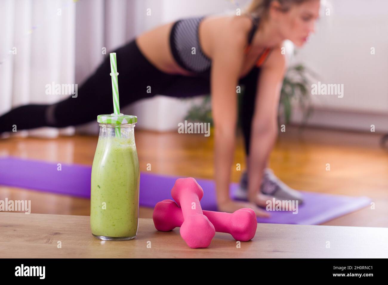 Primo piano di manubri e frullato in vaso con paglia e bella giovane donna stretching in background. Concetto di stile di vita sano Foto Stock