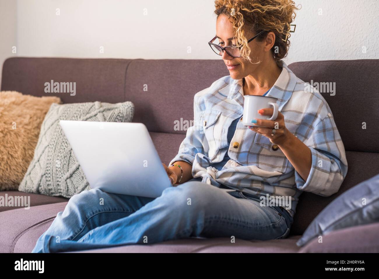 Sicuro felice adulto giovane donna utilizzare computer portatile seduto sul divano a casa in relax tempo libero attività bere caffè o tè e sorridere - moder Foto Stock