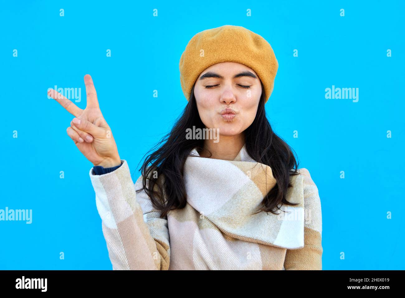 Foto di bella signora sognante soffio aria bacio spettacolo v-segno chiudere occhi indossare beretto beige e cappotto isolato biue colore sfondo Foto Stock