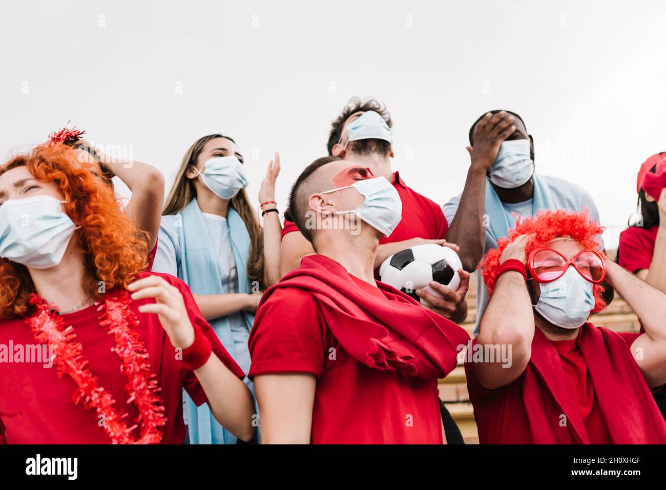 Frustrato gruppo di tifosi di calcio multirazziale in maschera protettiva che guarda la loro squadra di calcio perdere - Focus su uomo nel centro Foto Stock
