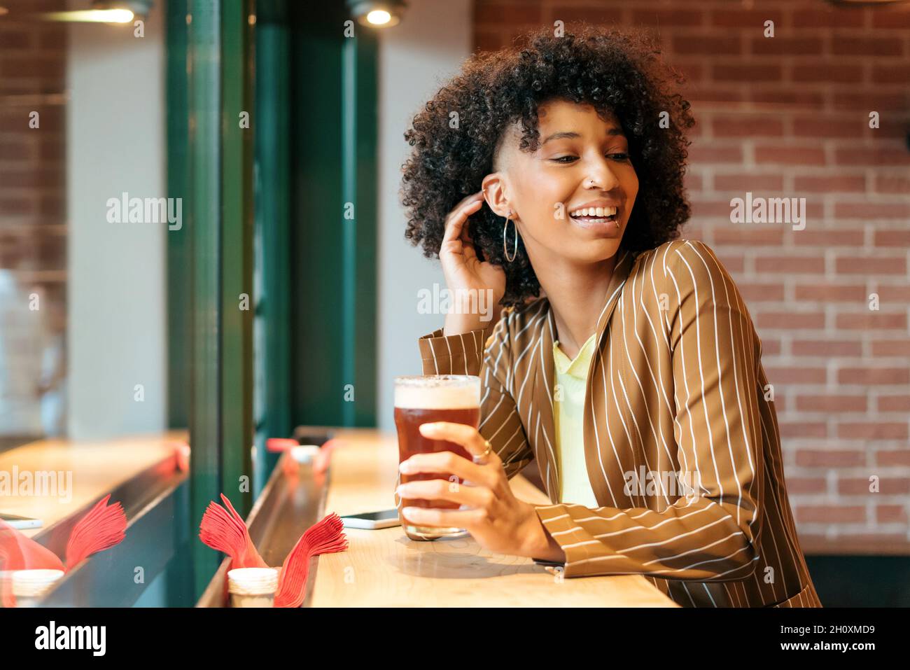 Vista laterale della giovane ragazza afro-americana allegra con eleganti capelli afro che indossano blazer a righe godendo un bicchiere di birra fredda mentre riposano al banco Foto Stock