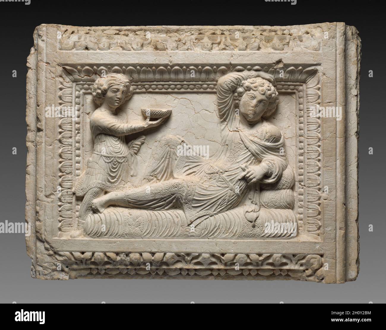 Rilievo tomba, 230. Siria, Palmyra, III secolo. Calcare; totale: 55.3 x 73.7 x 18.5 cm (21 3/4 x 29 x 7 5/16 poll.). Anche se il suo punto di ricerca è sconosciuto, questo rilievo alto quasi certamente una volta ha sigillato una nicchia sepolcrale di un mausoleo a Palmyra. Illustra un mix di tradizioni artistiche romane e partigiane. Adagiato all'interno di un sontuoso bordo di perle e foglie, un uomo ricco si risveglia su un letto ammortizzato di lusso mentre il suo assistente gli offre una ciotola di vino decorata con croccanti. Entrambi sono vestiti secondo la consuetudine Parthiana, indossando pantaloni e tuniche pieghettate, ma la posa languida dell'uomo è presa Foto Stock
