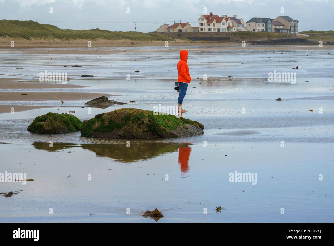 Una donna in giacca arancione che cammina sulla spiaggia a Rhosneigr, Anglesey, Galles, Regno Unito Foto Stock