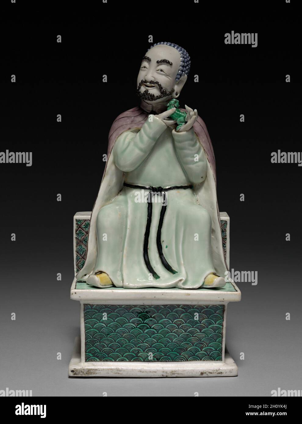 Lohan Holding a fu Dog, 1662-1722. Cina, provincia di Jiangxi, forni Jingdezhen, dinastia Qing (1644-1911), regno di Kangxi (1662-1722). Porcellana con decorazione in smalto vetrificato Famille verte; totale: 24.3 x 12.6 cm (9 9/16 x 4 15/16 poll.). Questo lohan, un discepolo di Buddha con poteri soprannaturali, era forse parte di un altare di casa o di un oggetto decorativo in una famiglia. Si dice che Lohans, noto anche come arhats, sia in grado di domare le tigri e calpestare l'acqua. Qui il lohan è mostrato come una figura monklike, tenendo un fu-dog.This lohan e la sua coppia che tiene la pesca d'immortalità (1964.191.1) incarnano un hu Foto Stock