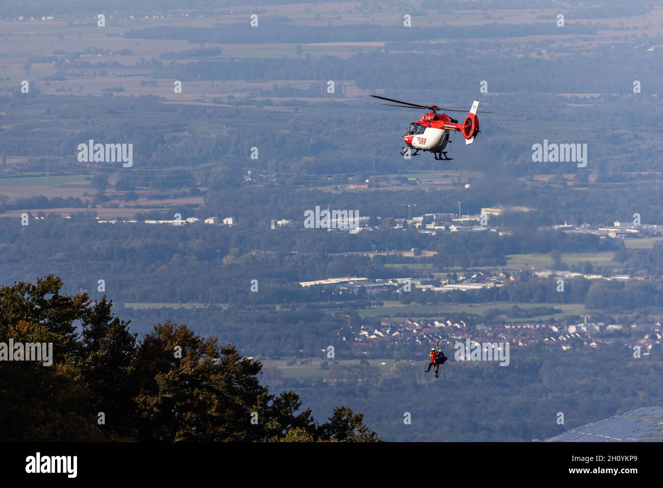 15 ottobre 2021, Baden-Wuerttemberg, Waldkirch: Un membro ciascuno del Bergwacht e del DRF Luftrettung appendere su una corda sotto un elicottero di salvataggio, mentre la valle del Reno può essere visto sullo sfondo. Durante l'addestramento del verricello, l'equipaggio della stazione di Friburgo della DRF Luftrettung (cartello di chiamata Christoph 54) pratica le operazioni con il verricello sotto un elicottero insieme al Black Forest Mountain Rescue Service. Questo verricello può essere utilizzato per salvare i pazienti da terreni impraticabili, nonché per trasportare medici e paramedici in regioni difficili da raggiungere. Foto: Philipp von Ditfurth/dpa Foto Stock