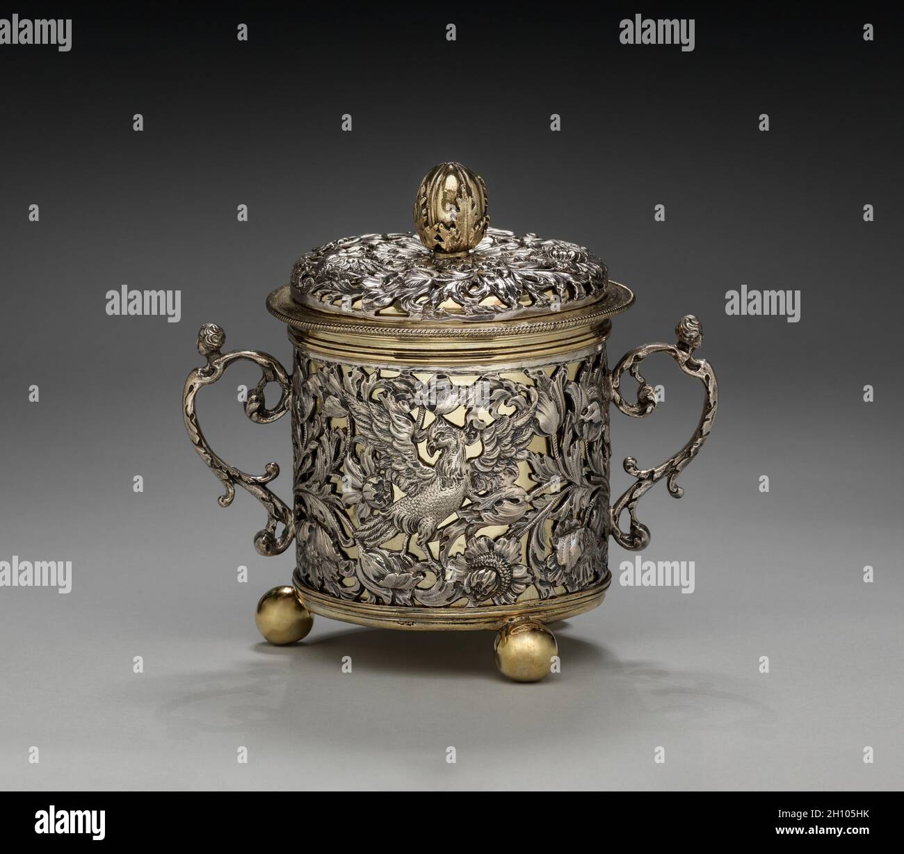 Tazza a due manine e coperchio, 1677. Inghilterra, Londra, XVII secolo (Carlo II). Argento e argento dorato; totale: 17.2 x 11.5 cm (6 3/4 x 4 1/2 pollici). A causa delle sue dimensioni e ornamento, questo recipiente può aver funzionato come una tazza caudle, contenente una miscela di birra o vino e pane, farinata d'avena, uova, zucchero e spezie che è stato dato alle donne dopo il parto per aiutarli a riguadagnare la loro forza. Il fogliame a scorrimento e gli uccelli esotici lungo l'esterno di questa tazza erano probabilmente basati su stampe circolanti nel 1600. Le figure femminili reclinabili che tappano le maniglie e il germoglio di acanto finiale sul coperchio recal Foto Stock