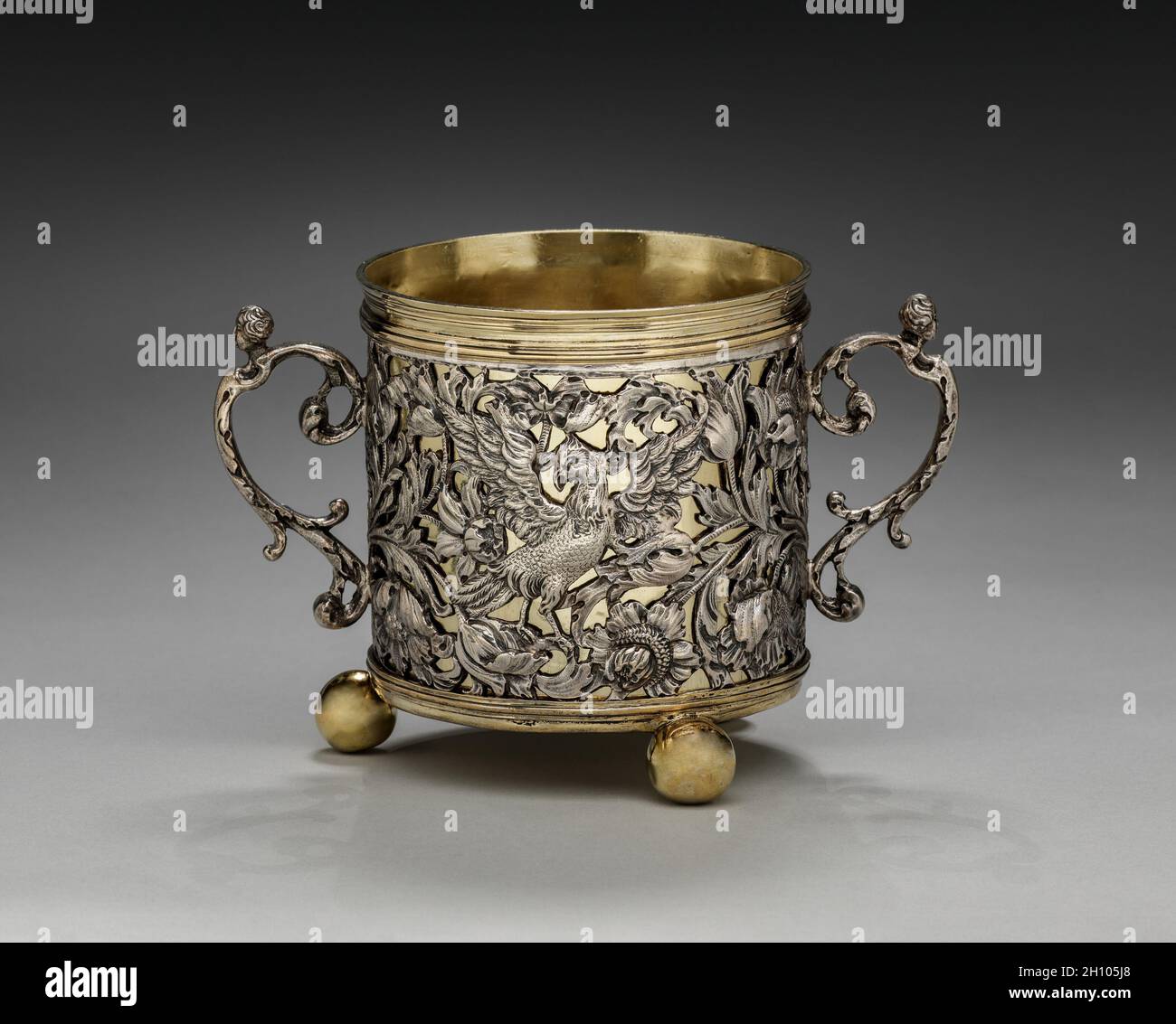 Tazza a due maneggiate, 1677. Inghilterra, Londra, XVII secolo (Carlo II). Argento e argento dorato; totale: 17.2 x 11.5 cm (6 3/4 x 4 1/2 pollici). A causa delle sue dimensioni e ornamento, questo vaso può aver funzionato come una tazza di caudle, contenente una miscela di birra o vino e pane, farinata d'avena, uova, zucchero e spezie che è stato dato alle donne dopo il parto per aiutarli a riguadagnare la loro forza. Il fogliame a scorrimento e gli uccelli esotici lungo l'esterno di questa tazza erano probabilmente basati su stampe circolanti nel 1600. Le figure femminili reclinabili che tappano le maniglie e l'acantus gemma finiale sul coperchio di accompagnamento re Foto Stock