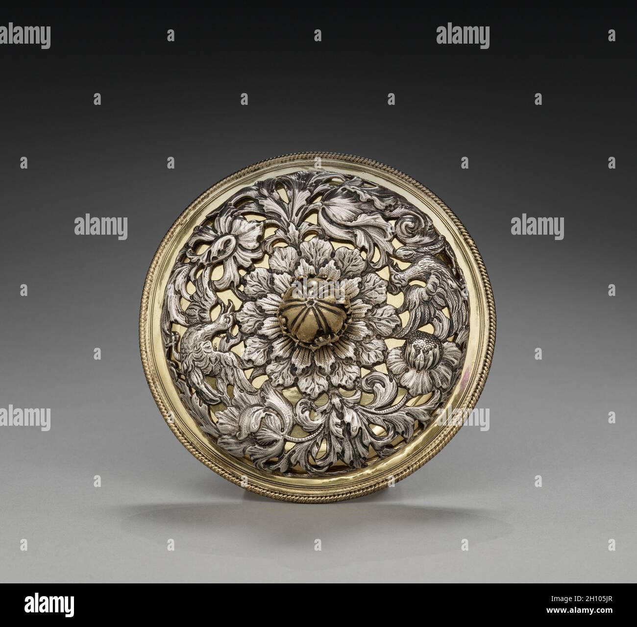 Coperchio per una tazza a due maneggiate, 1677. Inghilterra, Londra, XVII secolo (Carlo II). Argento e argento dorato; totale: 17.2 x 11.5 cm (6 3/4 x 4 1/2 pollici). A causa delle sue dimensioni e ornamento, il recipiente che accompagna questo coperchio può aver funzionato come una tazza caudle, contenente una miscela di birra o vino e pane, farinata d'avena, uova, zucchero e spezie che è stato dato alle donne dopo il parto per aiutarli a riguadagnare la loro forza. Il fogliame a scorrimento e gli uccelli esotici lungo l'esterno della tazza erano probabilmente basati su stampe circolanti nel 1600. Le figure femminili reclinabili che tappano le maniglie e il germoglio di acanto f Foto Stock