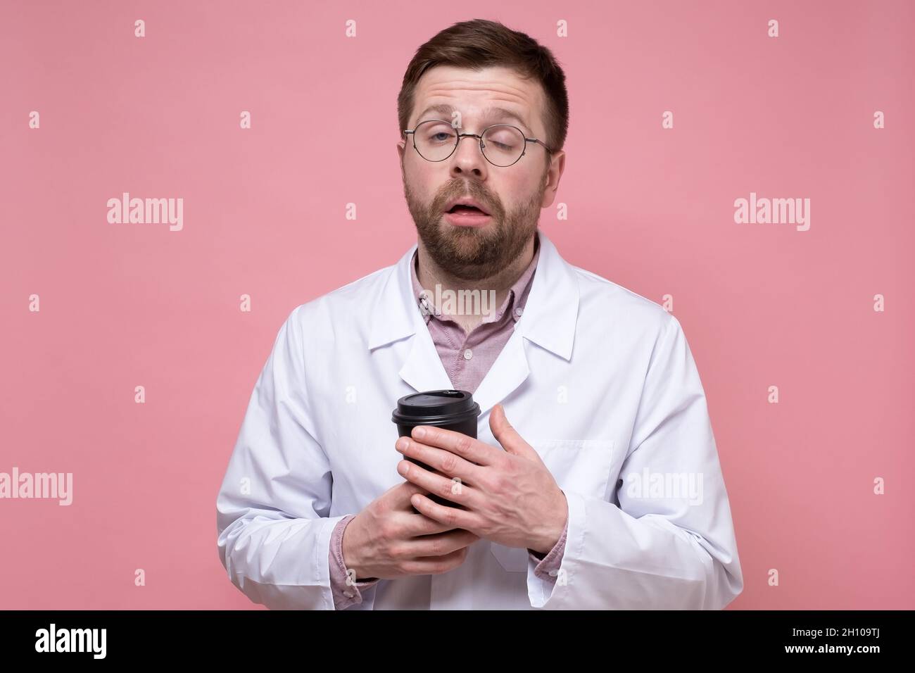 Medico in un cappotto bianco e bicchieri è molto stanco e sembra assonnato, tiene una tazza di carta di caffè nelle mani e cerca di aprire gli occhi. Foto Stock