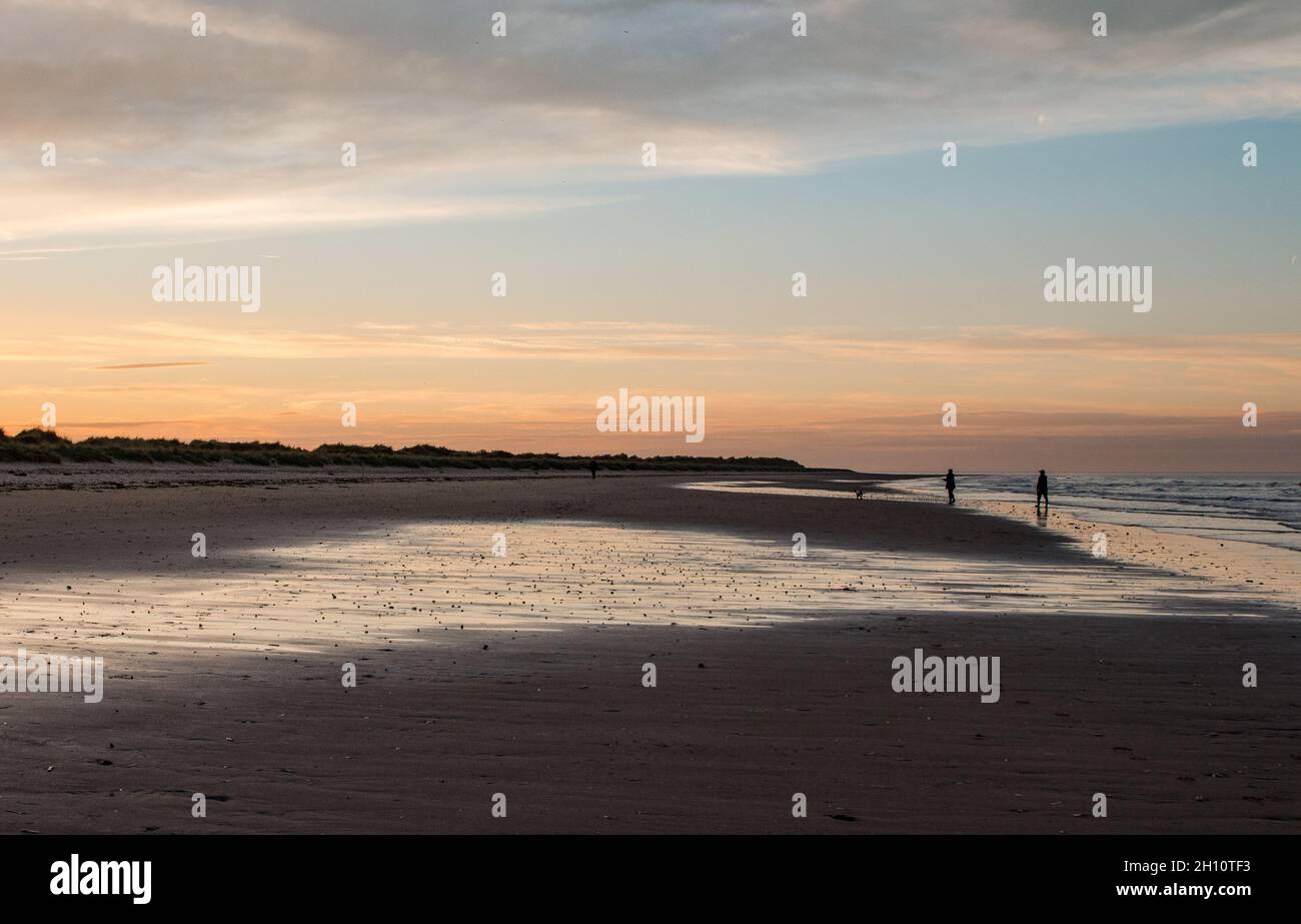 Figure ombreggiate su una spiaggia di Norfolk con onde e riflessi acquosi nella luce sbiadente del tramonto. Foto Stock