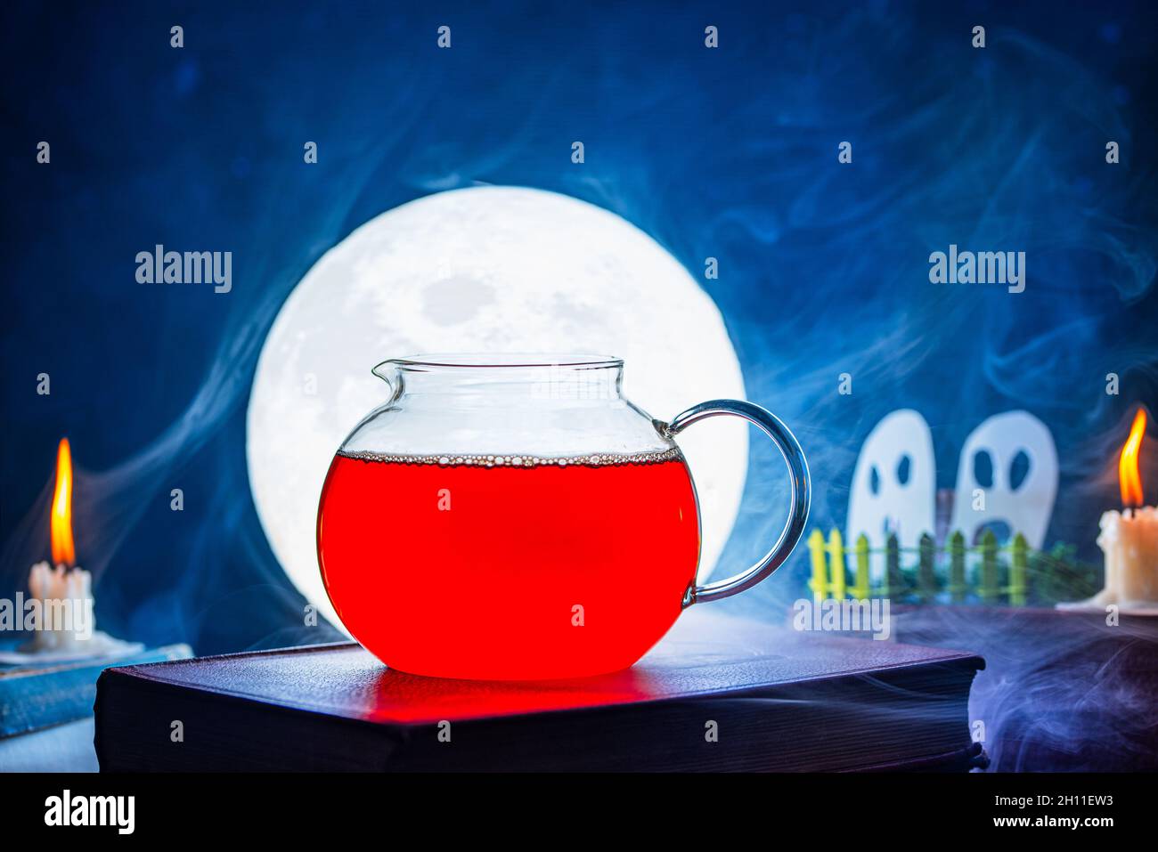 tè in una caraffa, sulla parte anteriore della luna che brilla brillantemente, tra due candele che sono accese e hanno una fiamma arancione contro un backgr blu Foto Stock